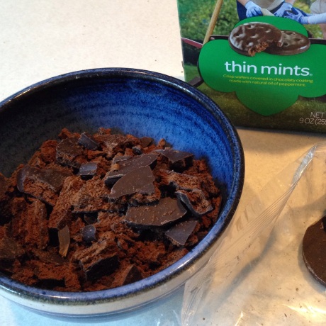 Pieces of Thin Mint cookies sit in a blue bowl, with a box of the cookies in the background.